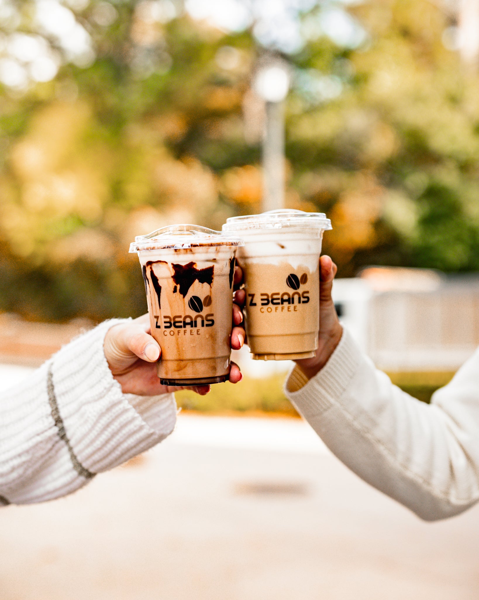 Two hands holding iced coffee cups with 'Z Beans Coffee' branding outdoors.