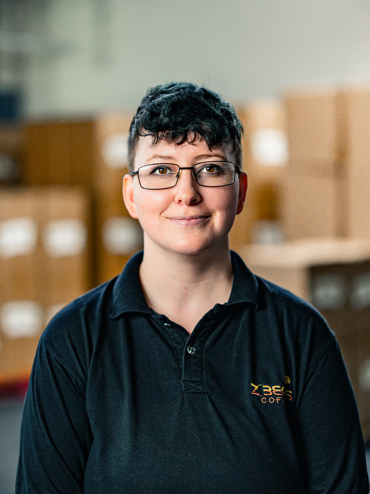 Woman wearing a black Z Beans polo shirt with a logo in an indoor setting with cardboard boxes.