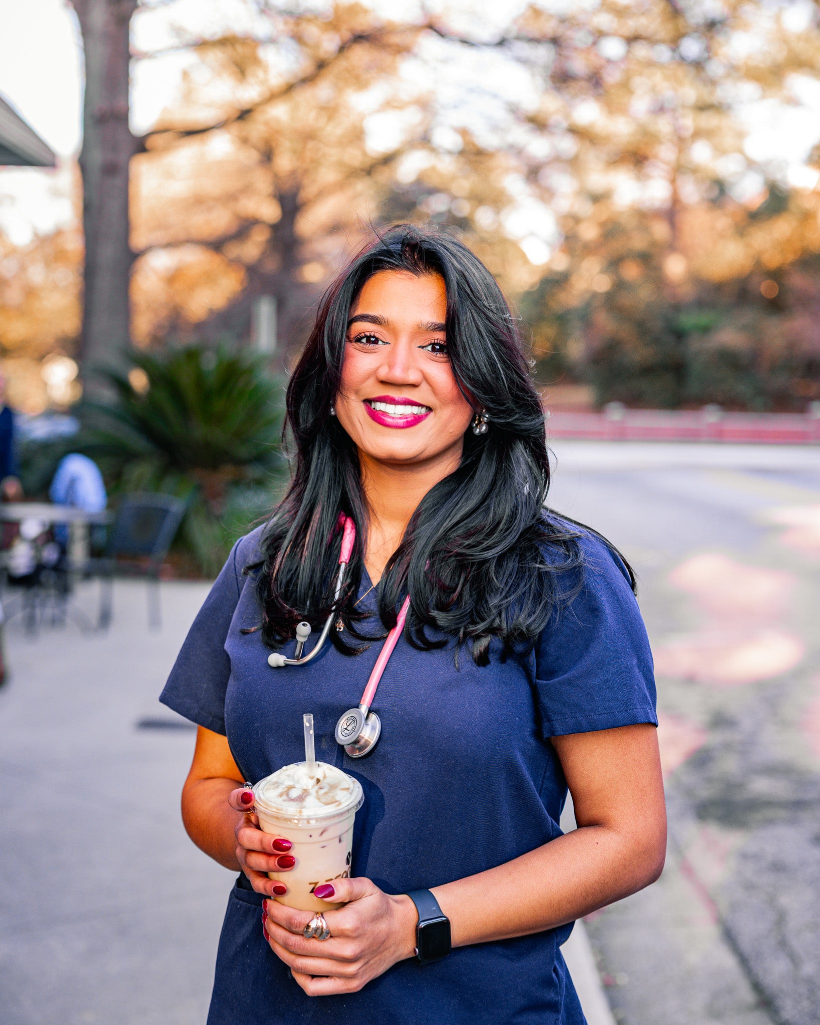 Person in blue scrubs holding a drink outdoors with trees in the background