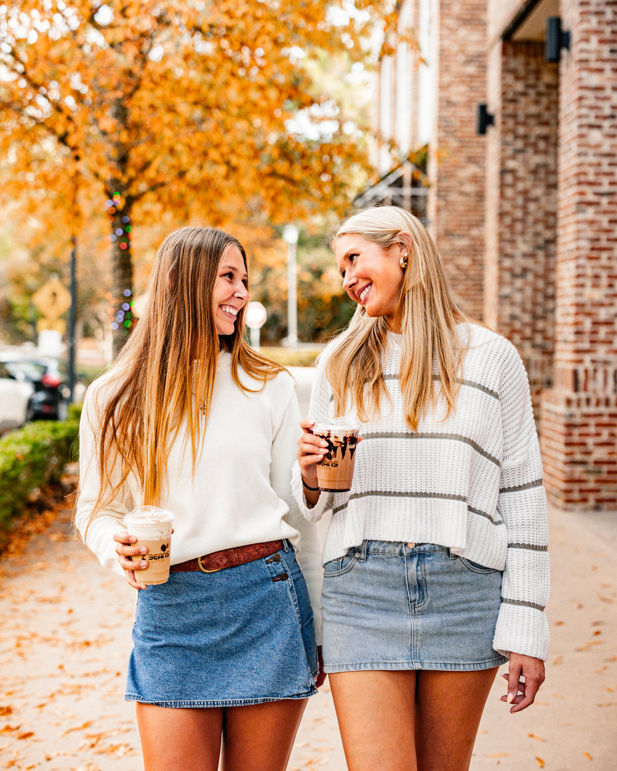 Two women walking outdoors in autumn, holding coffee cups smiling at eacj other.