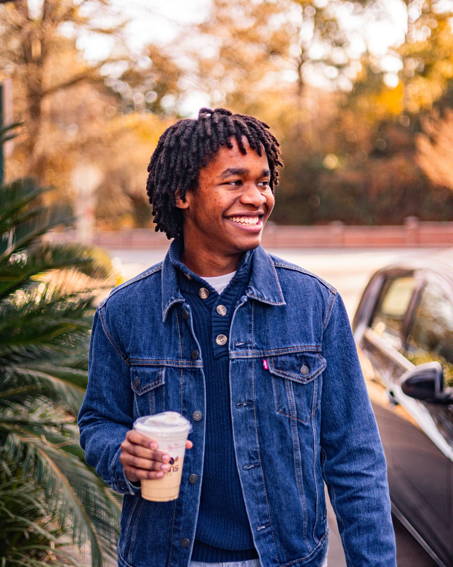 Man in a denim jacket holding a coffee cup outdoors with trees and car in the background