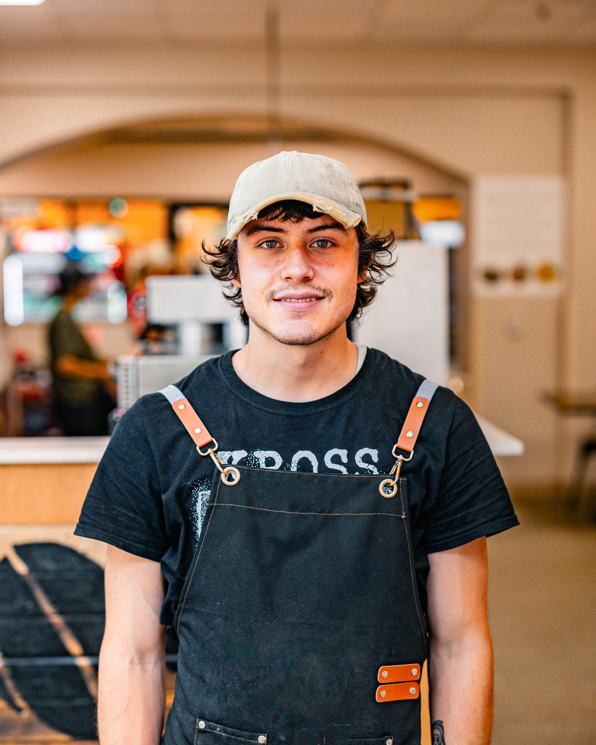 Man wearing a black t-shirt with 'BOSS' text and an apron in an indoor cafe setting.