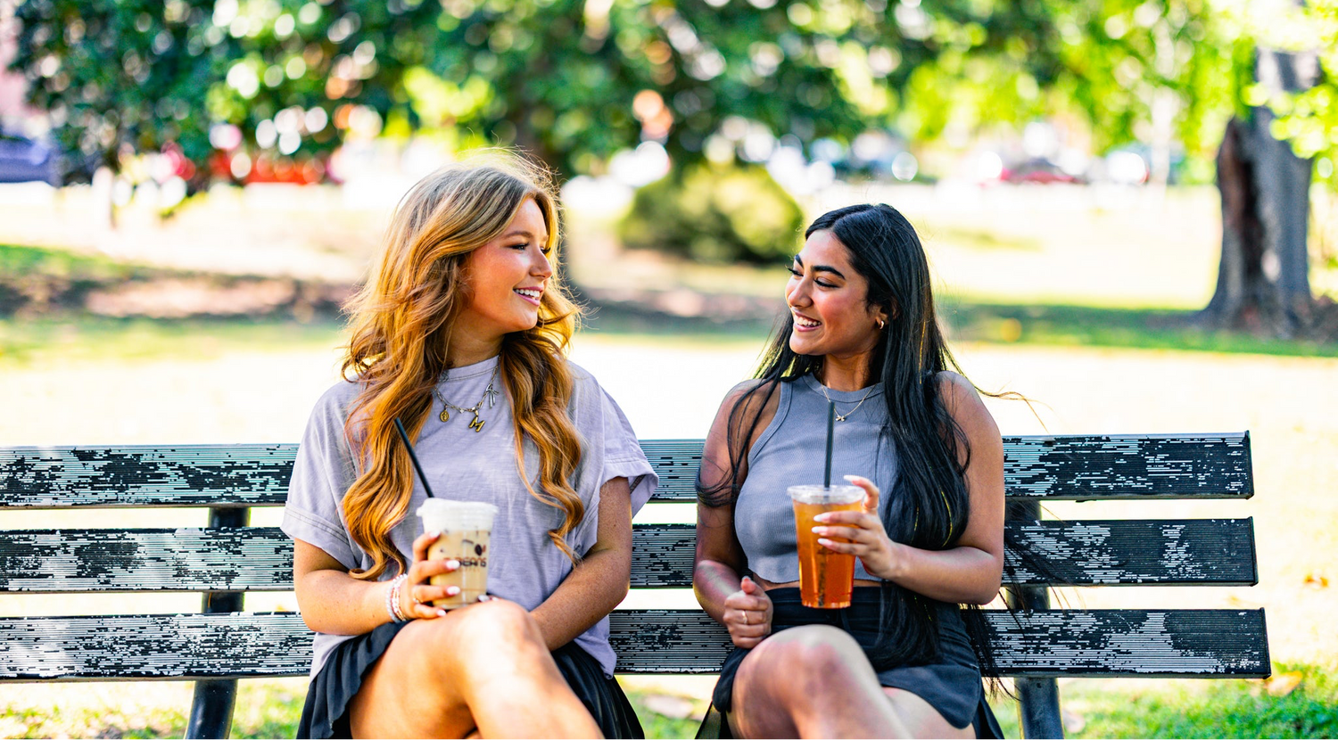 Two women sitting on a park bench, one with a coffee cup and the other with a tea, surrounded by greenery.