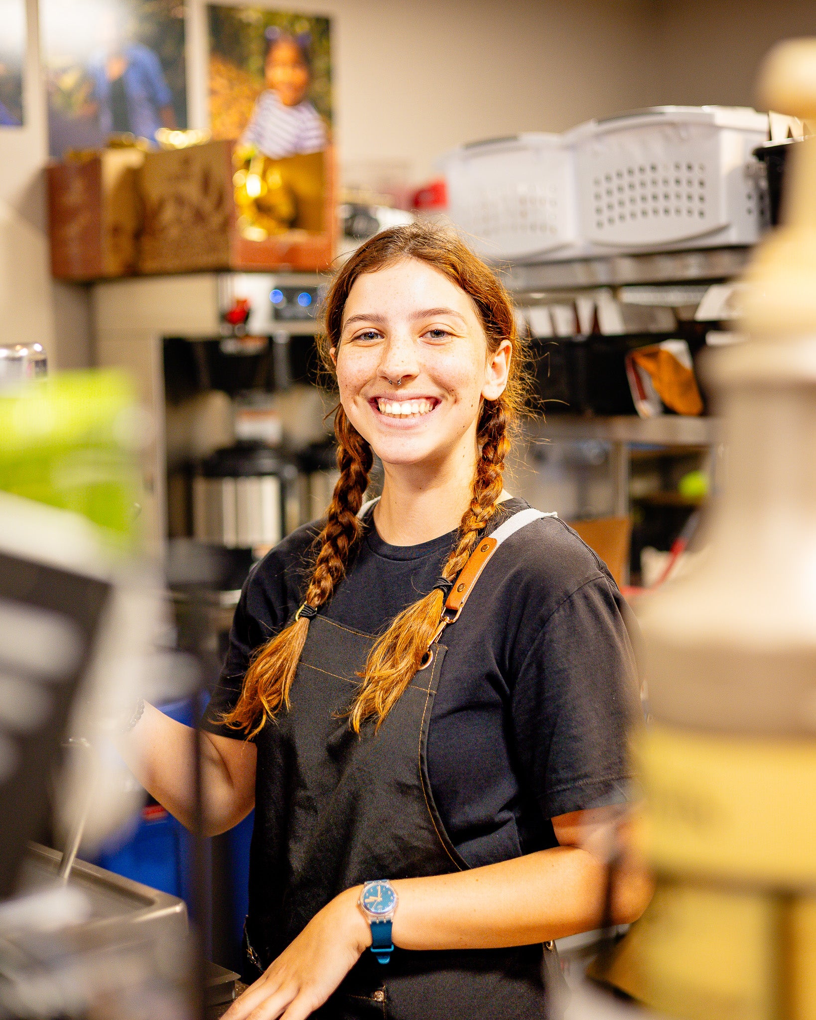 Person with braided hair wearing a black shirt and apron in a cafe counter setting.