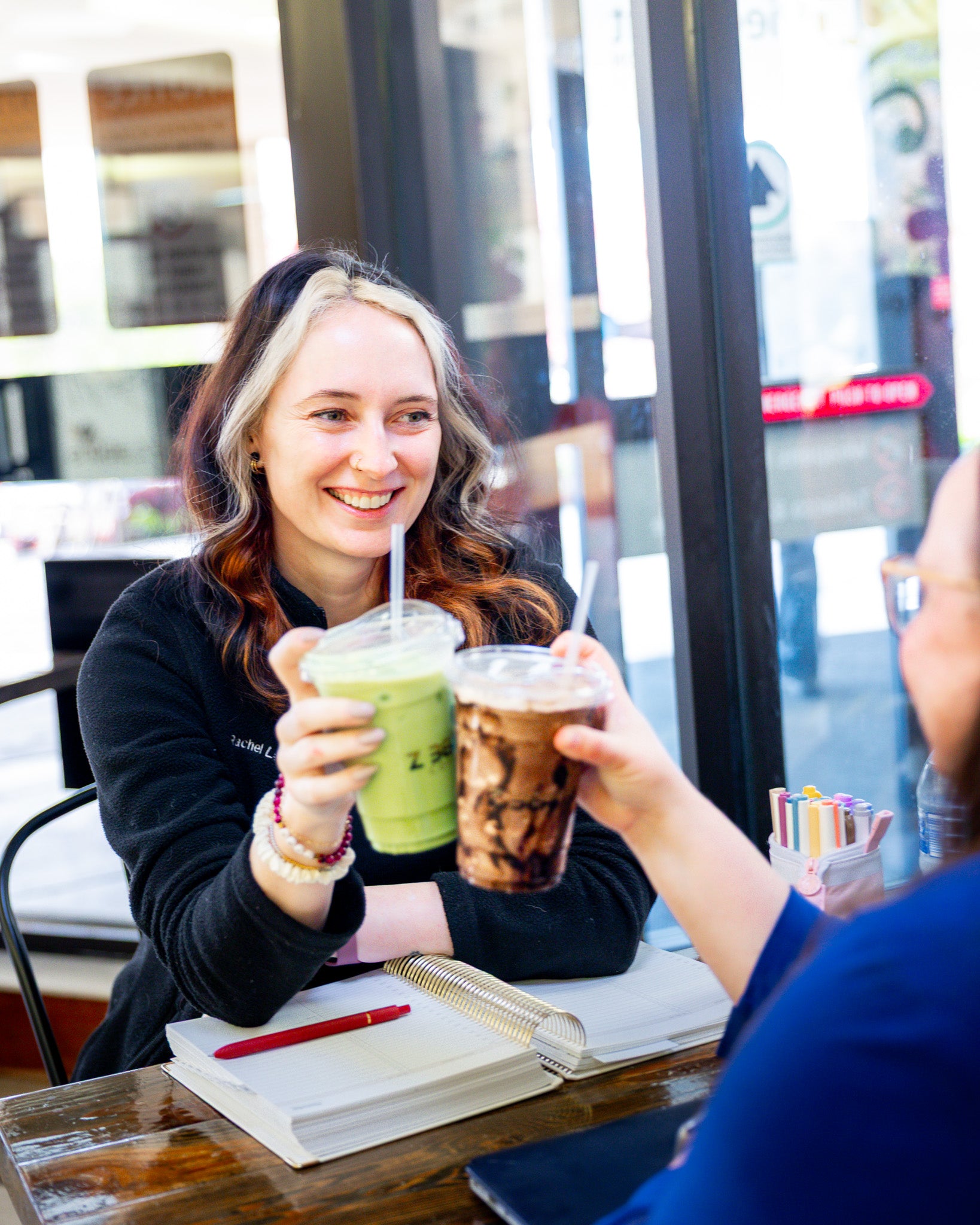 Two people at a cafe, one holding a green smoothie and the other a iced coffee, with a cityscape in the background.