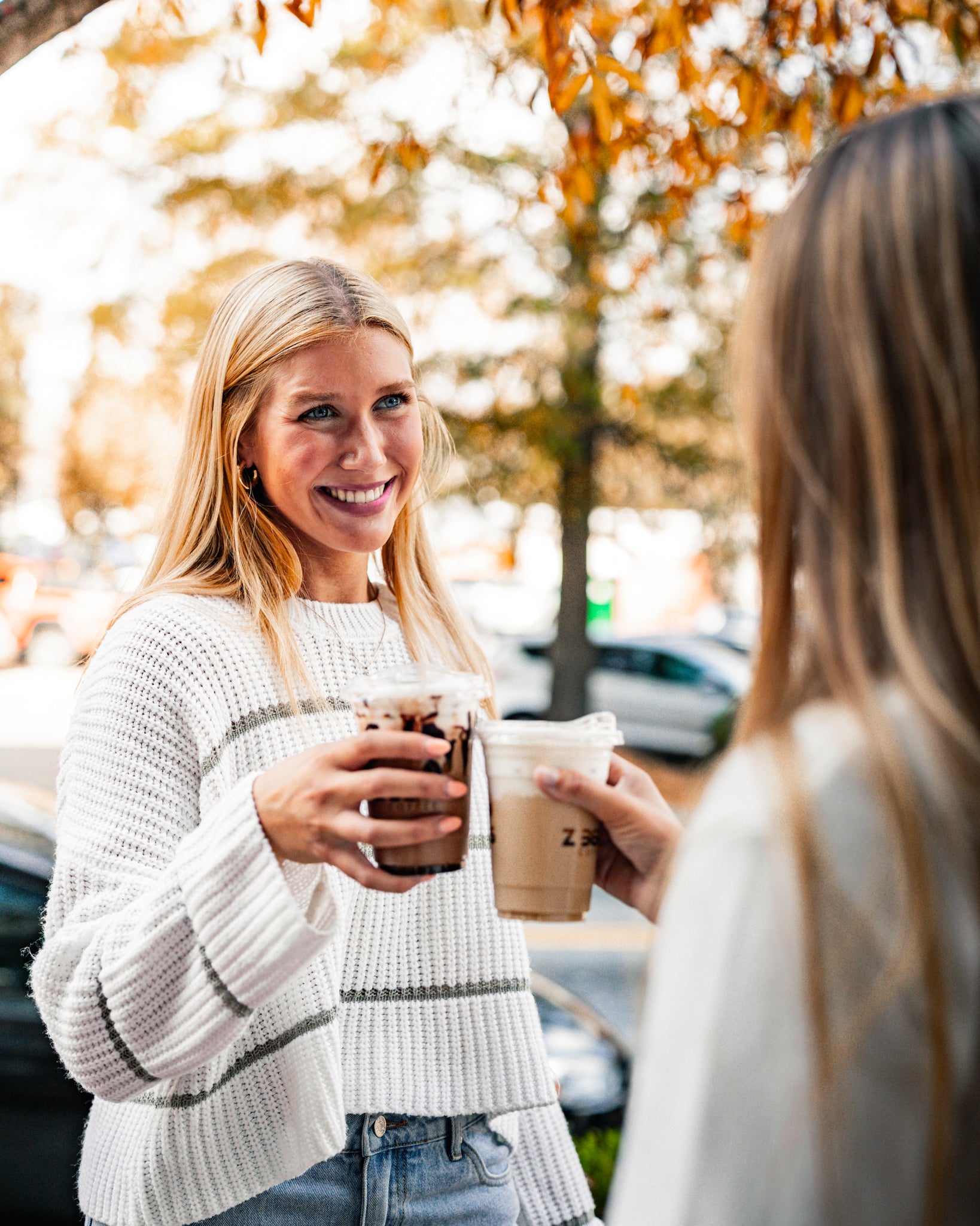 Two women holding coffee cups outdoors with trees and cars in the background